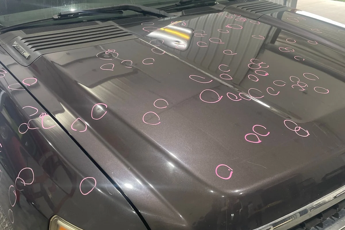 A truck hood with dozens of hail dents individually circled in pink marker, the shop bay door standing open behind it.
