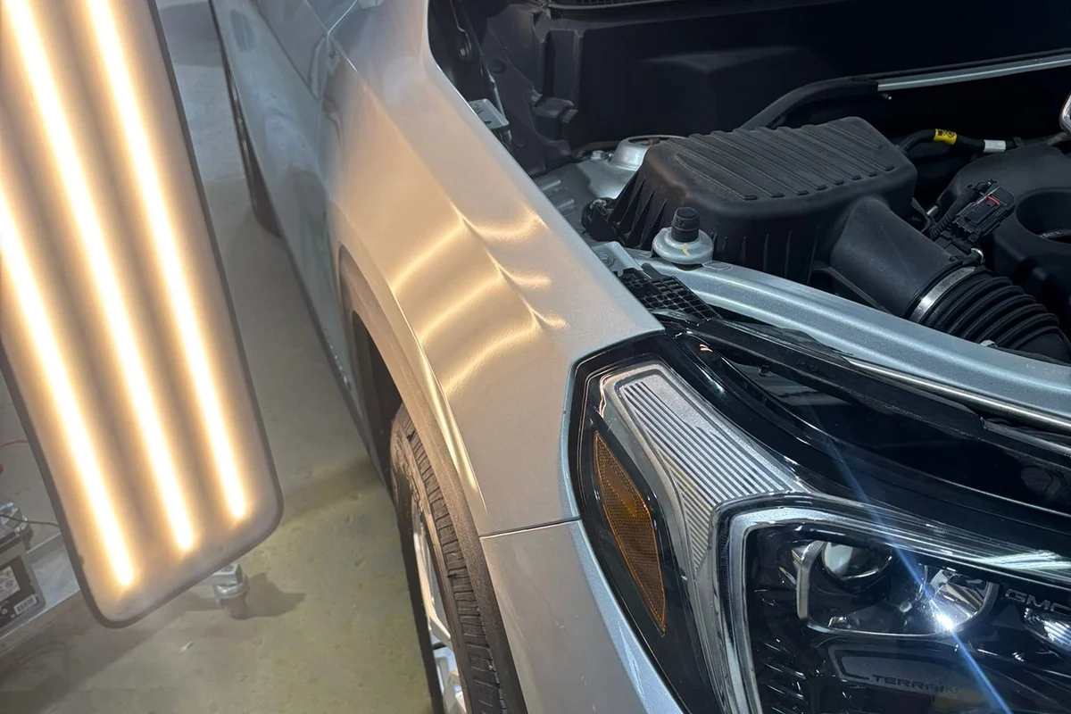 An LED line board on a stand throwing banded light across a vehicle fender, the bands bending where hail dents sit below the surrounding panel.