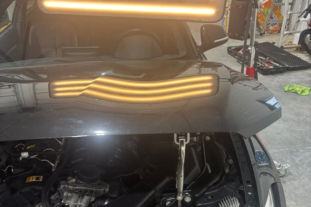A dark car with its hood raised, a line board on a rolling stand throwing banded light across the hood to reveal hail dents.