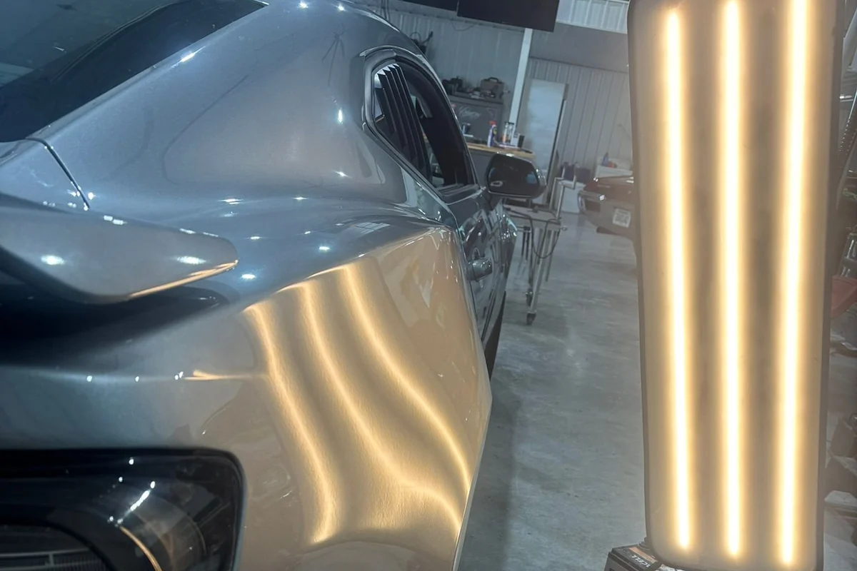 An LED line board on a rolling stand beside a vehicle rear quarter panel, its banded reflection bending across a field of hail dents.