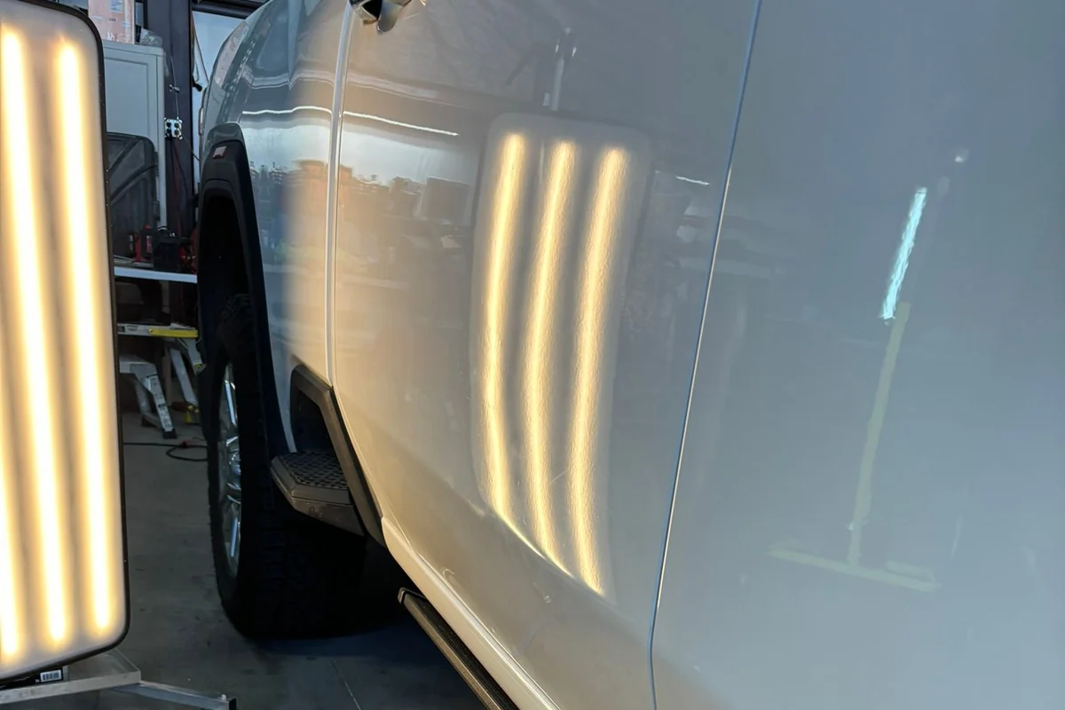 A freestanding LED line board lighting the door and rocker of a truck in the shop bay, its banded reflection rippling across the panel.