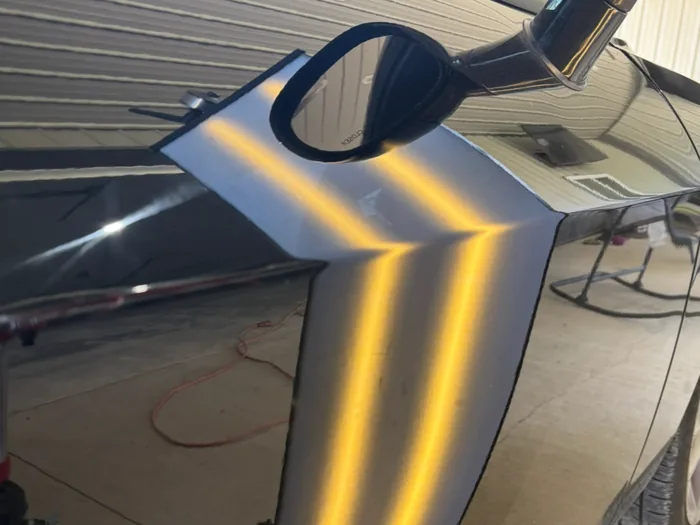 An LED line board on a rolling stand casting gold bands across a black car door, revealing the hail dents in the panel.